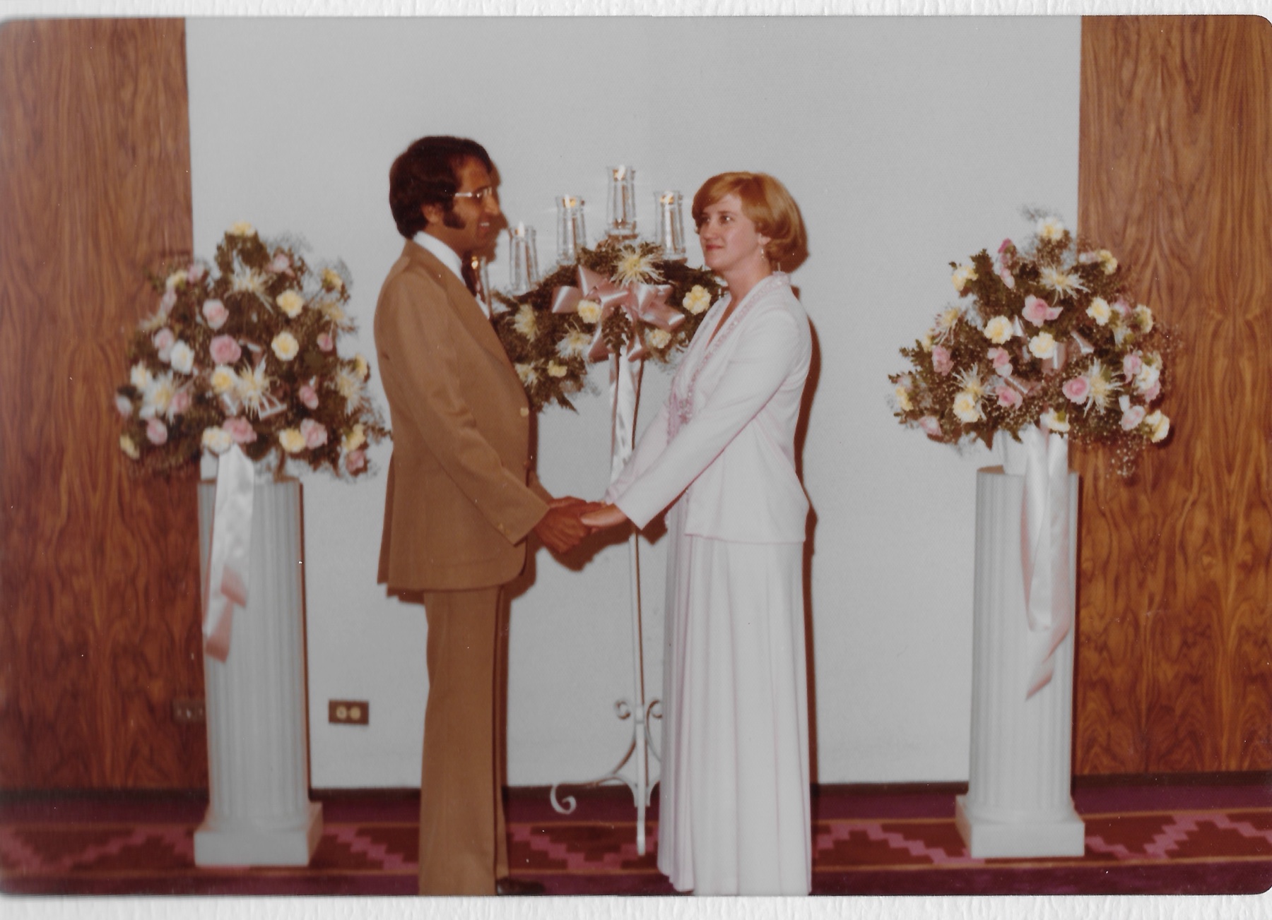 Mary Lee and Madhu holding hands at the altar between two tall stands of white flowers